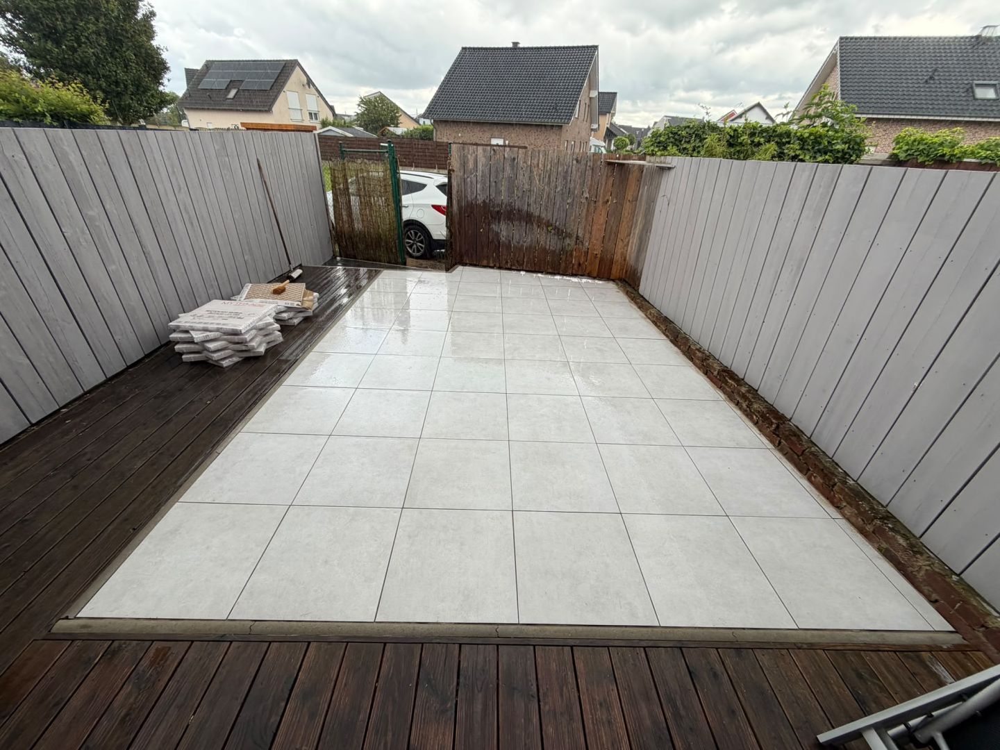 Helle Terrassenplatten neben einem dunklen Holzbelag vor einem grauen Zaun